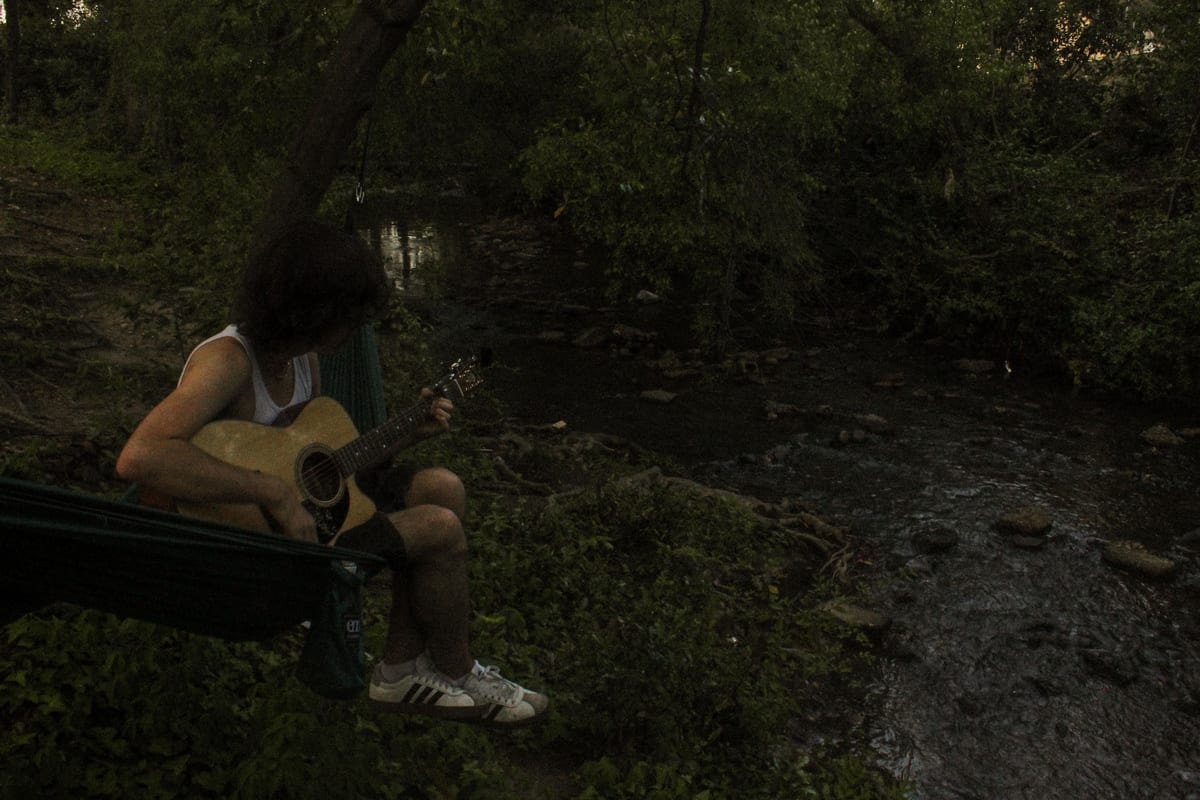 Figure seated in a hammock beside a creek at dusk, playing an acoustic guitar